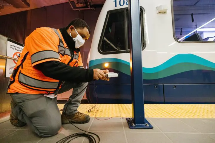 Technician in orange safety vest and mask works on a train platform's electrical components.
