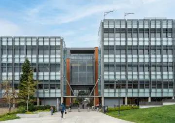 Modern glass and metal building facade with people walking on a campus-like pathway below a blue sky.