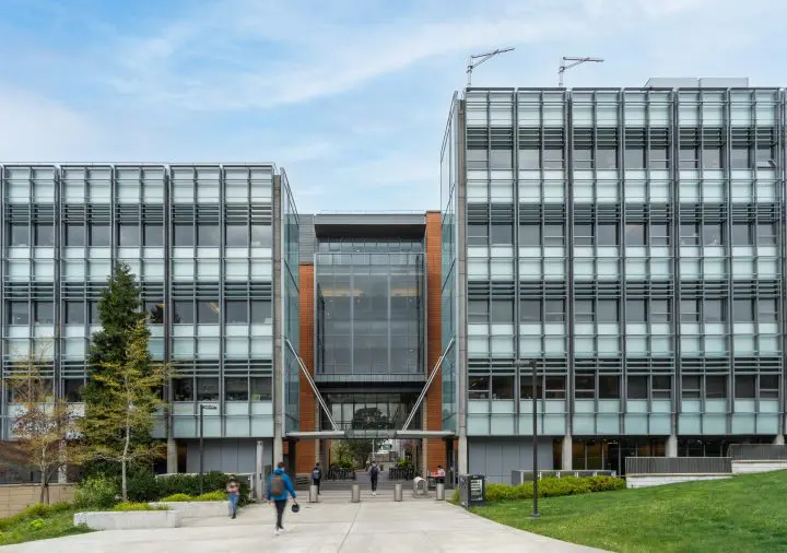Modern glass and metal building facade with people walking on a campus-like pathway below a blue sky.