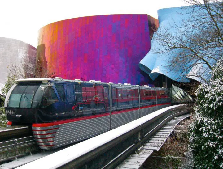 Monorail travels past architecturally striking colorful building in winter setting, amidst snow-dusted trees and tracks.