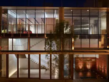 Modern glass building facade at night, showcasing sleek interior spaces and staircases with warm lighting.