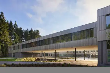 Modern building with expansive glass and metal facade, surrounded by trees. A person walks below on a paved path.