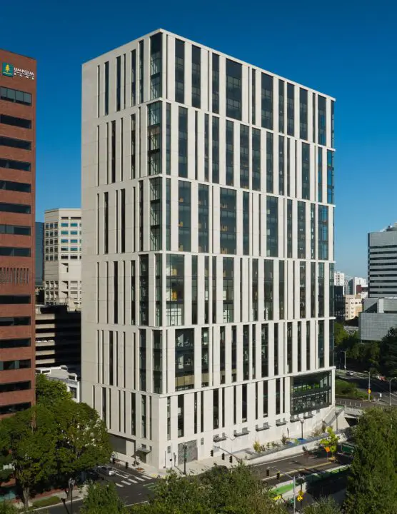 Modern high-rise building under clear blue sky, surrounded by urban landscape and trees, reflecting contemporary architecture.