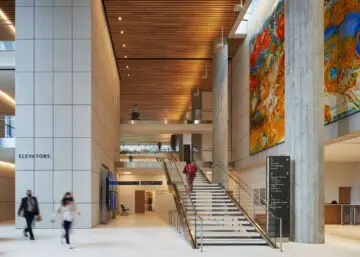 Modern building interior with large colorful artwork, stairs, and people walking. Bright lighting and spacious design.