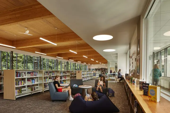 Open, modern library with people reading and studying in a cozy seating area, surrounded by bookshelves and large windows.