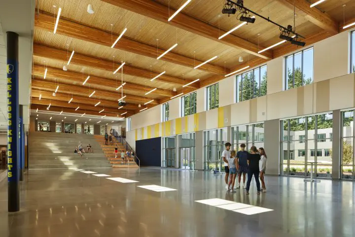Modern school atrium with high ceilings and large windows, featuring socializing students and tiered seating. Bright and spacious.