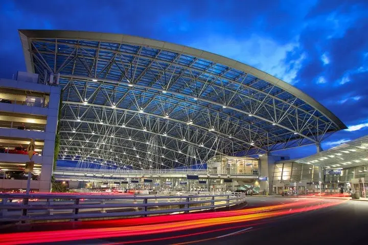Modern airport entrance lit at dusk, showcasing architectural design with bright blue sky and motion blur of car lights.