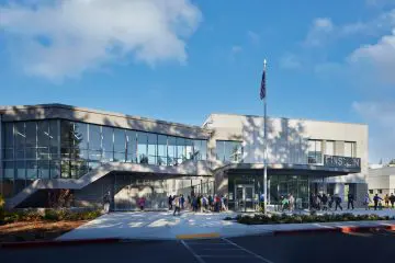 Modern school building with large windows, flagpole, and people outside on a sunny day. Blue sky with fluffy clouds.