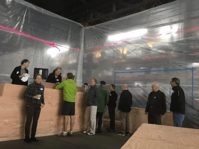 Group of people wait in line at an indoor counter, interacting with staff. Meeting or event in a warehouse-like setting. Casual attire.