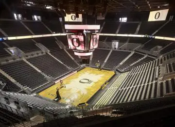 Empty basketball arena with a large central screen, logo on the court, and tiered seating, ready for a thrilling game.