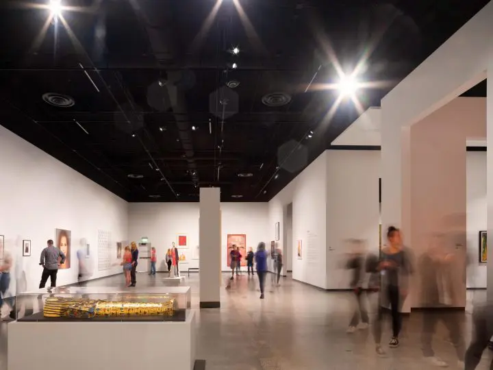 Modern art gallery interior with blurred visitors viewing exhibits, bright overhead lighting, and contemporary artwork displayed on walls.