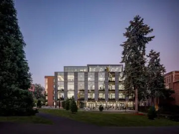 Modern glass building at dusk, surrounded by tall trees, with warm interior lighting. Peaceful campus setting.
