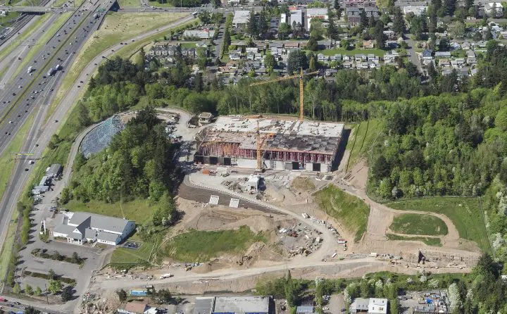 Aerial view of a construction site with cranes, surrounded by trees and near a highway and residential area.