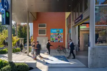 Busy urban station exterior with people walking, vibrant poster on brick wall, bike rack, and greenery.