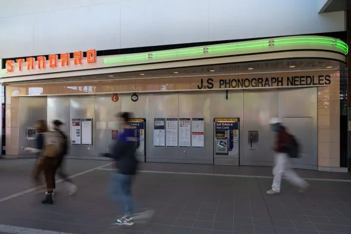 Blurred commuters walk past a station ticket kiosk under a retro neon sign with green and red lights.