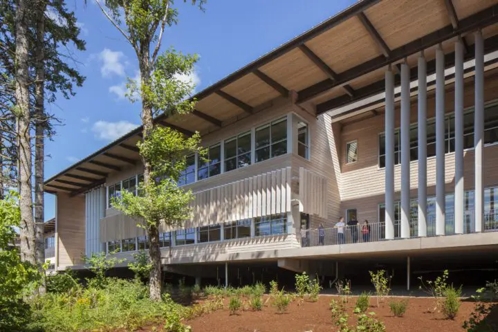 Modern wooden building with large windows and outdoor balcony, surrounded by lush greenery under a clear blue sky.