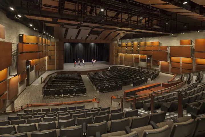 Spacious theater with performers rehearsing on stage; modern design, empty seats, warm lighting, and acoustic panels create an elegant ambiance.