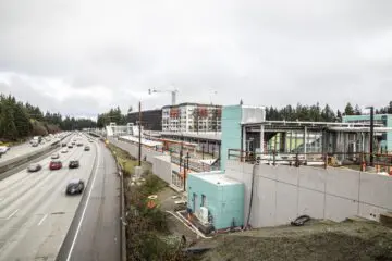 Highway beside a construction site with buildings and crane under a cloudy sky, surrounded by trees.