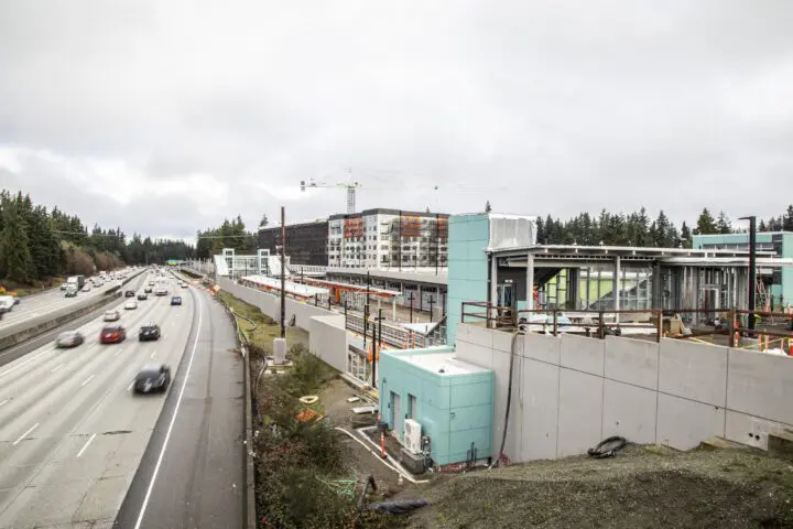Highway beside a construction site with buildings and crane under a cloudy sky, surrounded by trees.