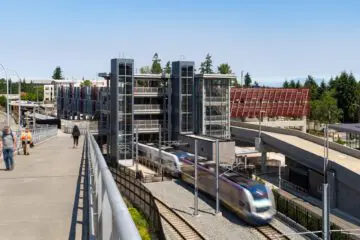 Modern light rail station with sleek design, moving train, and pedestrians in bright sunlight, surrounded by trees and clear sky.