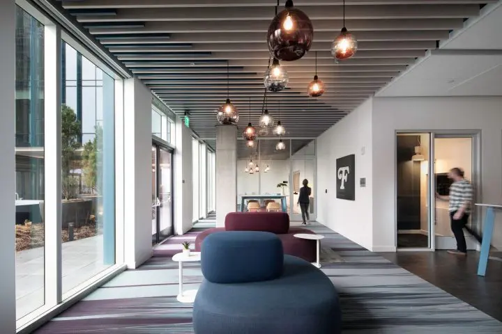 Modern office lounge with colorful seating, pendant lights, and large windows. Two people walking through the bright space.