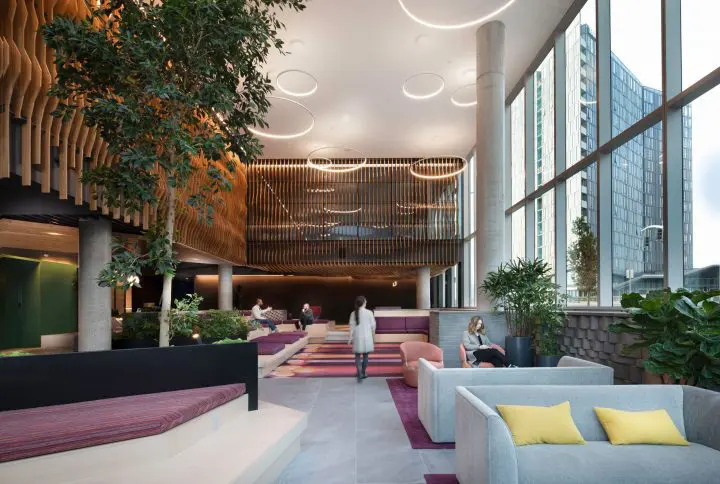Modern lobby with lush greenery, stylish seating, and large windows. People relax and converse in a bright, open space.
