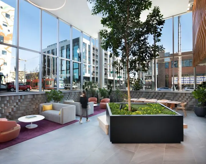 Modern office lobby with green indoor plants, seating, and large windows showcasing urban street view; person walking through.