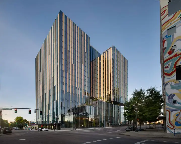 Modern glass skyscraper against a clear sky at an urban intersection, with colorful mural and trees in the foreground.