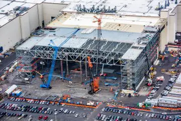 Aerial view of a construction site with cranes and heavy machinery building a large industrial structure with a parking lot nearby.