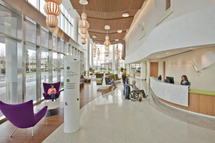 Modern children's hospital lobby with high ceilings, reception desk, seating area, and signage for elevators.