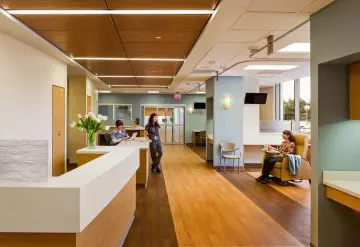 Modern hospital waiting area with reception desk, comfortable seating, and natural lighting. People sitting and working.