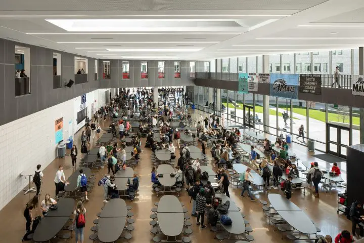 Spacious school cafeteria bustling with diverse students during lunch break, featuring long tables and large windows.