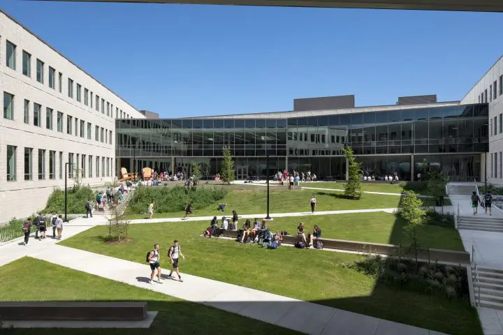 Modern campus courtyard with people walking and sitting on grass, surrounded by a glass-paneled building under a clear blue sky.