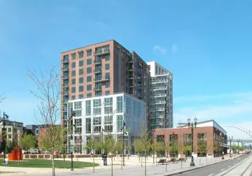 Modern urban apartment building with glass facade, trees, and blue sky. City street view with benches and green space.