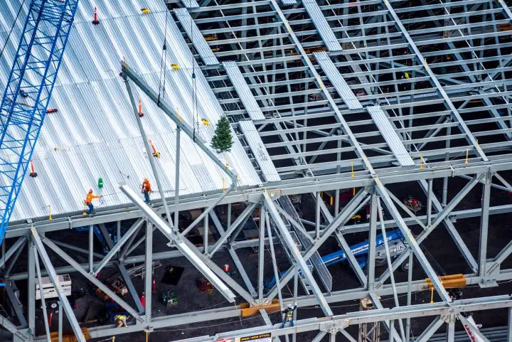 Construction workers on a steel frame structure, crane in background. A small tree visible, indicating progress milestone.