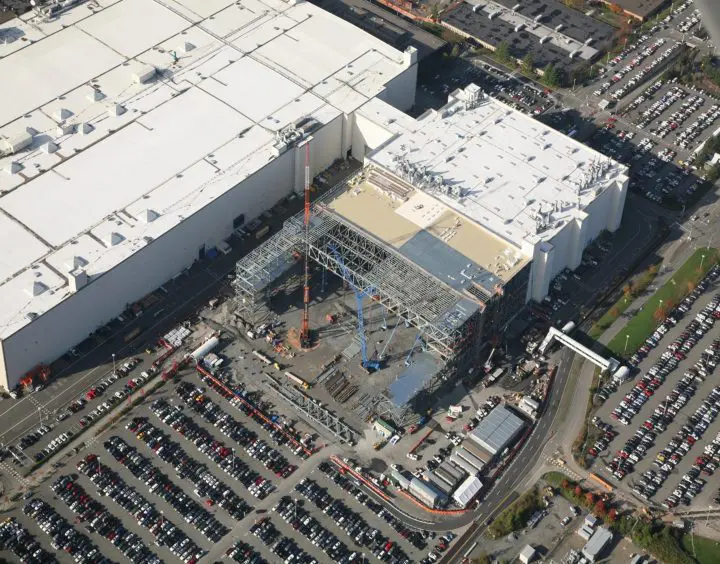 Aerial view of a large construction site next to an industrial building, with cranes and a busy parking lot in the foreground.