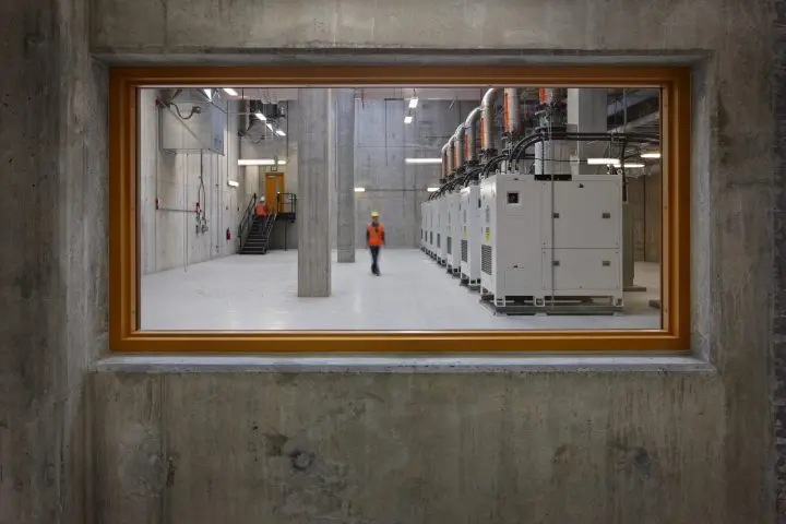 Industrial facility interior with machinery and a worker in orange vest and hard hat, viewed through a large window frame.