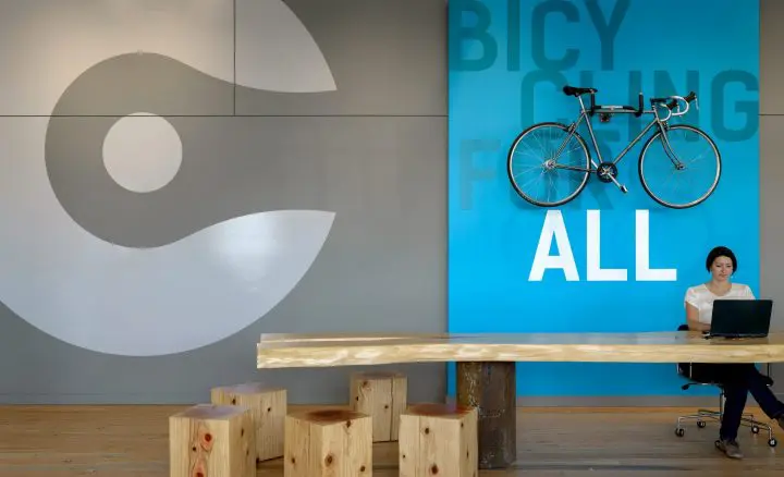 Modern office space with bike on wall, wooden seating, and person working at a laptop. “Bicycling for all” slogan displayed.