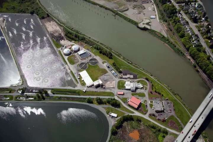 Aerial view of a large wastewater treatment plant between a river and a highway, showcasing settlement ponds and surrounding facilities.