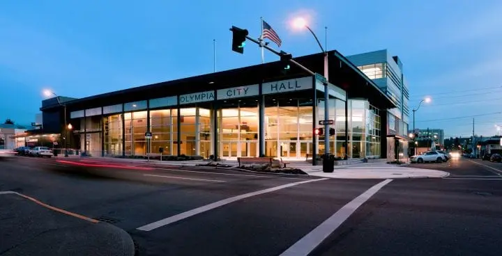 Modern city hall building with large glass windows at dusk, American flag flying, cars passing by on a well-lit street corner.