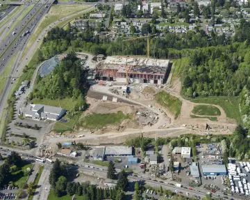 Aerial view of a large construction site near a highway surrounded by trees and buildings in a suburban area.