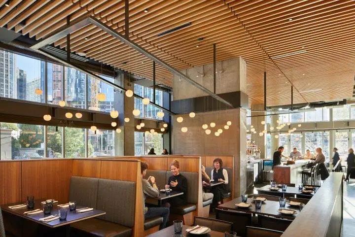 Modern restaurant interior with natural light, wooden decor, and patrons enjoying meals in cozy booths and at the bar.