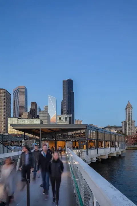 People walking on a modern pier with a scenic cityscape and skyscrapers in the background during sunset.