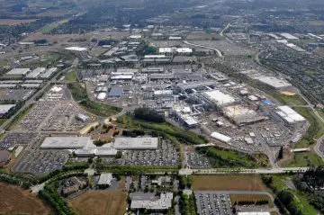 Aerial view of a large industrial complex surrounded by parking lots, roads, and greenery in a suburban landscape.