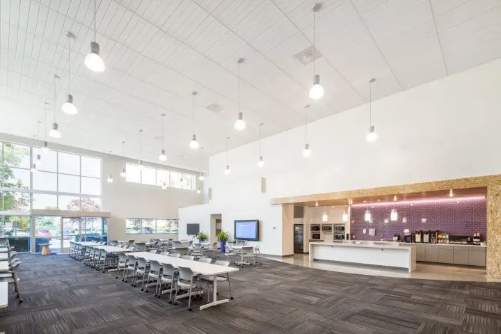 Modern open-plan office space with large windows, ample lighting, seating area, and kitchen amenities. Bright and spacious design.