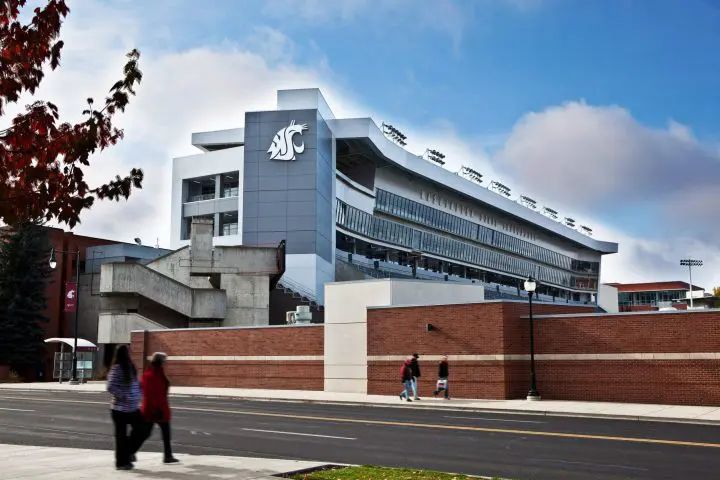 Modern university stadium exterior with people walking; Washington State University logo visible. Bright day with blue sky.