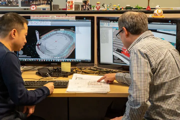 Two individuals collaborate on 3D design at a workstation with dual monitors displaying architectural software and sketches.