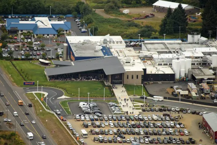 Aerial view of a large dairy processing plant and visitor center with parking lot, surrounded by trees and roads.