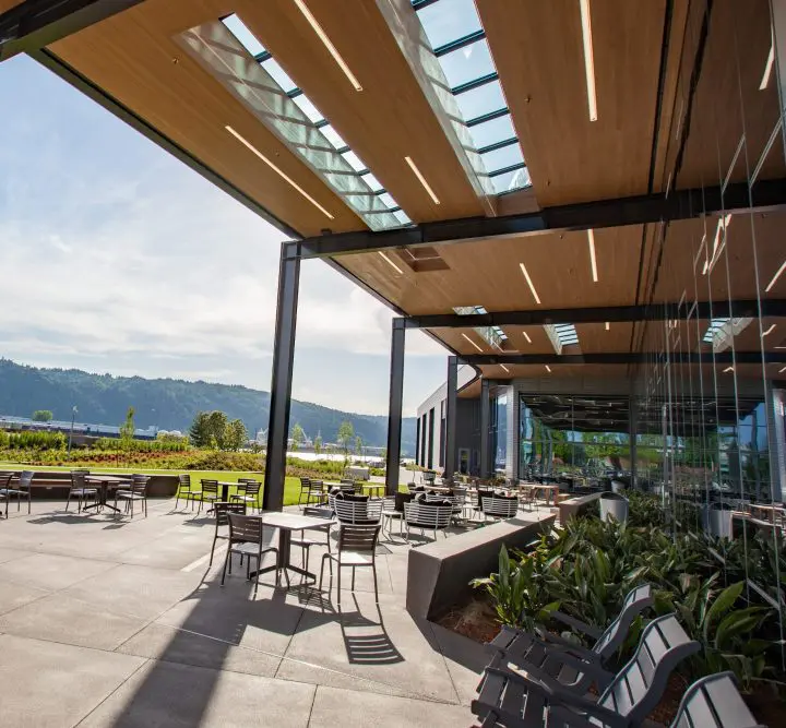 Modern outdoor patio with tables, chairs, and greenery under a wooden ceiling. Mountains and clear skies in the background.