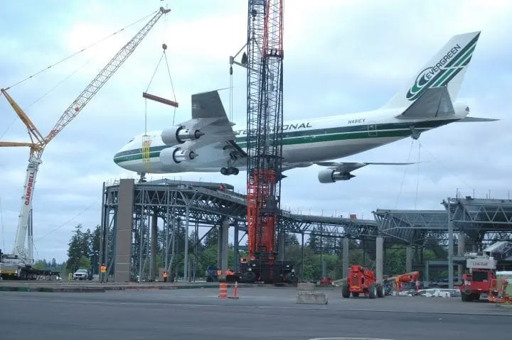 Airplane being lifted by cranes during construction at an outdoor site, near a partially built platform, with cloudy sky.